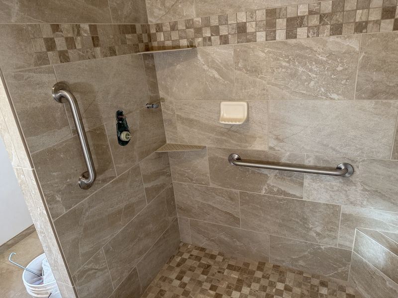 Accessible shower in travertine-look tile with grab bars and a pebble-tile floor.